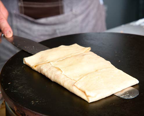 Bei einem mobilen Catering-Event von Reicherts benutzt eine Person einen Spatel, um einen rechteckigen Crêpe auf einer flachen, runden Grillplatte zuzubereiten und zu falten. Der Crêpe erscheint golden und leicht knusprig auf der Oberfläche.