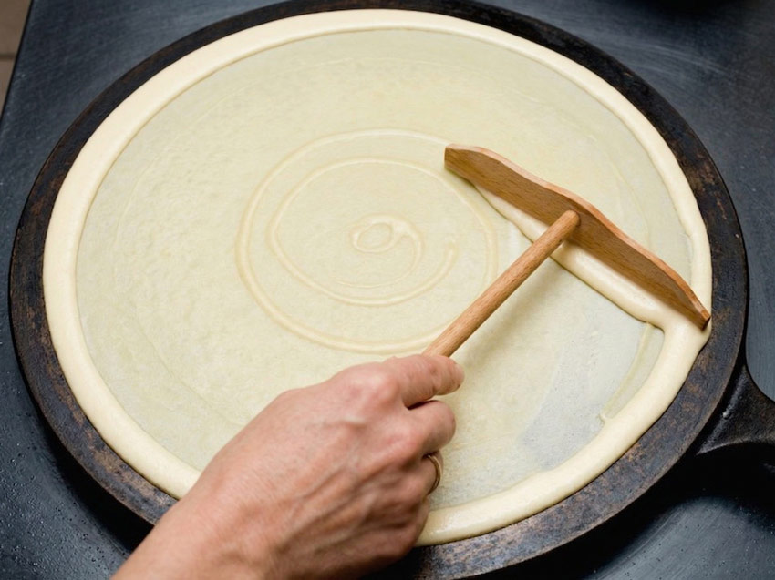 Bei einer reicherts-Veranstaltung verteilt eine Hand mit einem hölzernen Spachtel den Crêpeteig gleichmäßig in einer kreisförmigen Bewegung auf einer runden, flachen, schwarzen Grillplatte - perfekt für das mobile Catering.